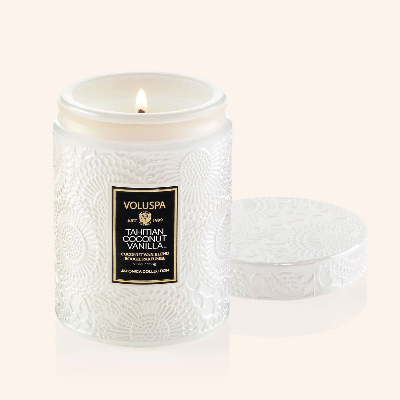 A glass candle with beautiful etched details sits in the middle of image.  Candle label reads "Voluspa Tahitian Coconut Vanilla"