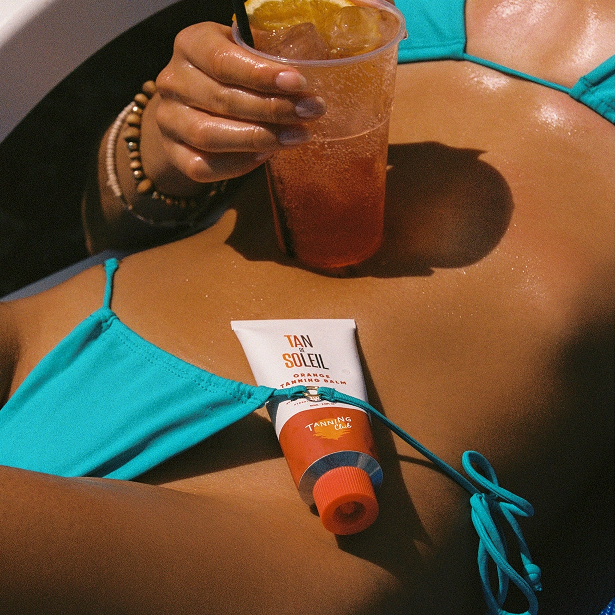 Person in turquoise bikini holding a drink with Tan Soleil tanning balm on arm