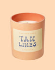 An apricot colored glass candle with a tan label, blue text that reads, 'Tan Lines'. 