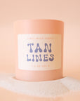 An  apricot colored glass candle with a tan label, blue text that reads, 'Tan Lines'. 