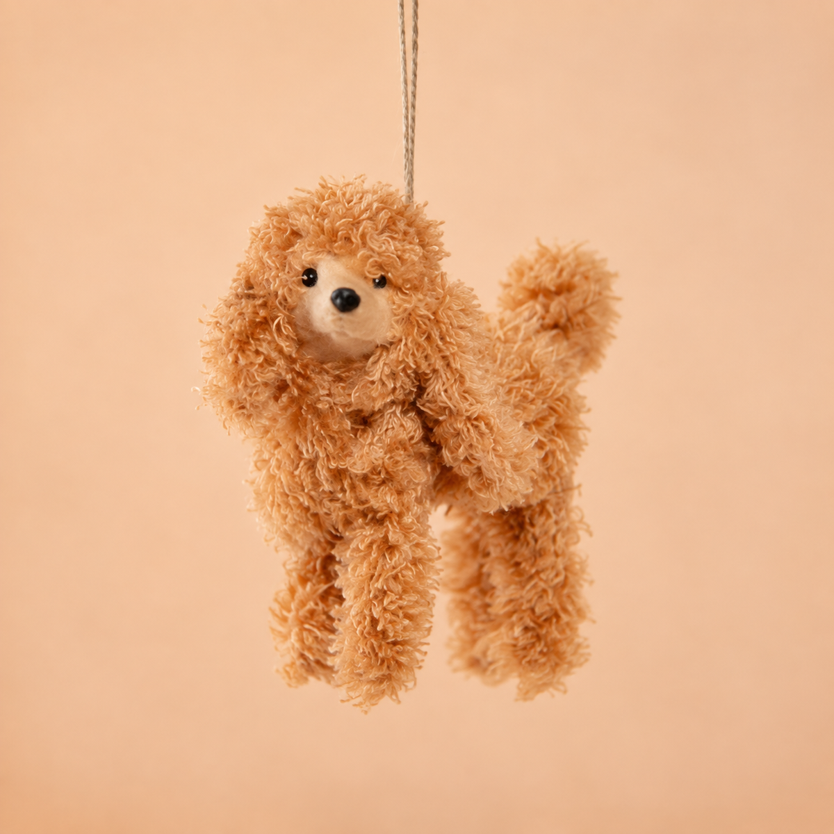 A small poodle ornament hanging from a thin string against a solid warm peach background.