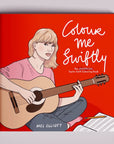 On a grey background is a red book with an illustration of Taylor Swift with a guitar and the white title on the right that reads, "Colour me Swiftly The Unofficial Taylor Swift Colouring Book". 