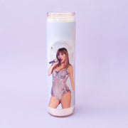 On a purple background is a prayer candle featuring a light blue background and the iconic Taylor Swift on the front in a sparkly onesie.