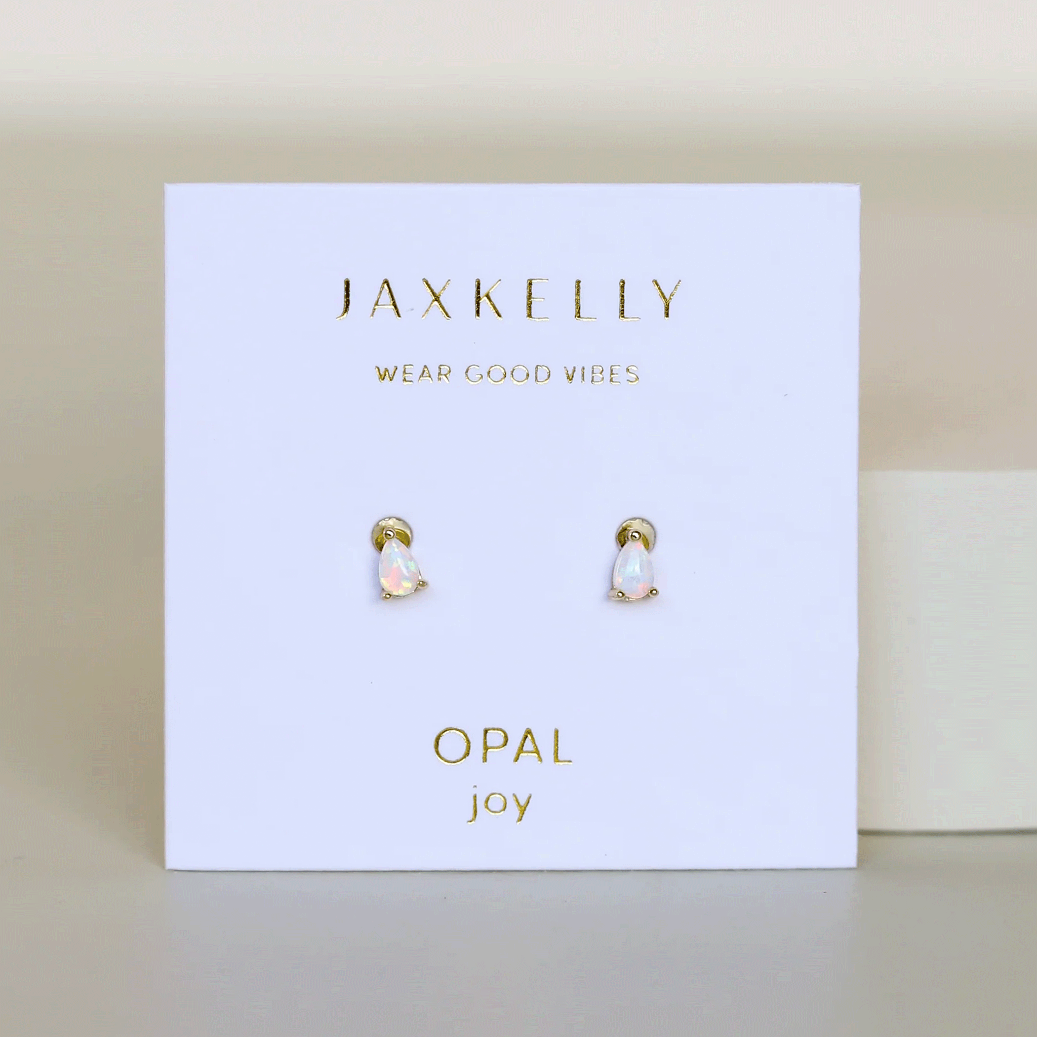 On a cream background is a small white cardboard packaging holding a pair of teardrop shaped white opal stud earrings. 