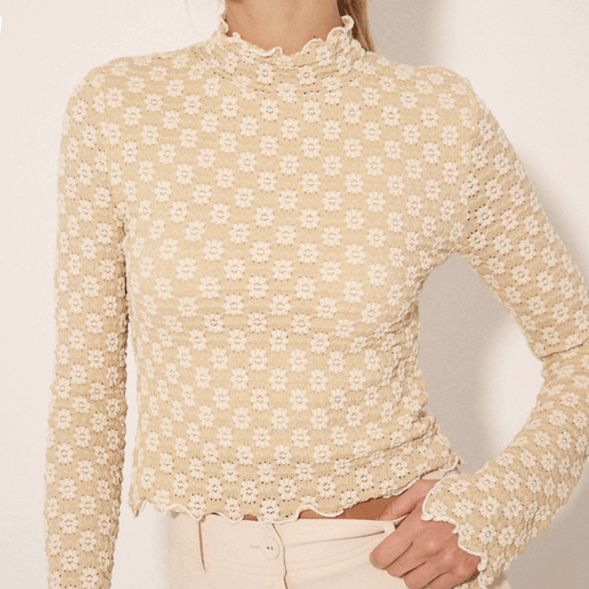 A model wearing a textured ivory top with a floral design and a mock neck that has a subtle ruffle detail.