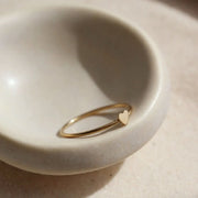 In a neutral ceramic bowl is a dainty gold ring with a tiny heart in the center. 