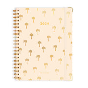 On a white background is a spiral bound planner with a light peachy color and a small gold foiled palm tree design as well as "2024" in the center. 