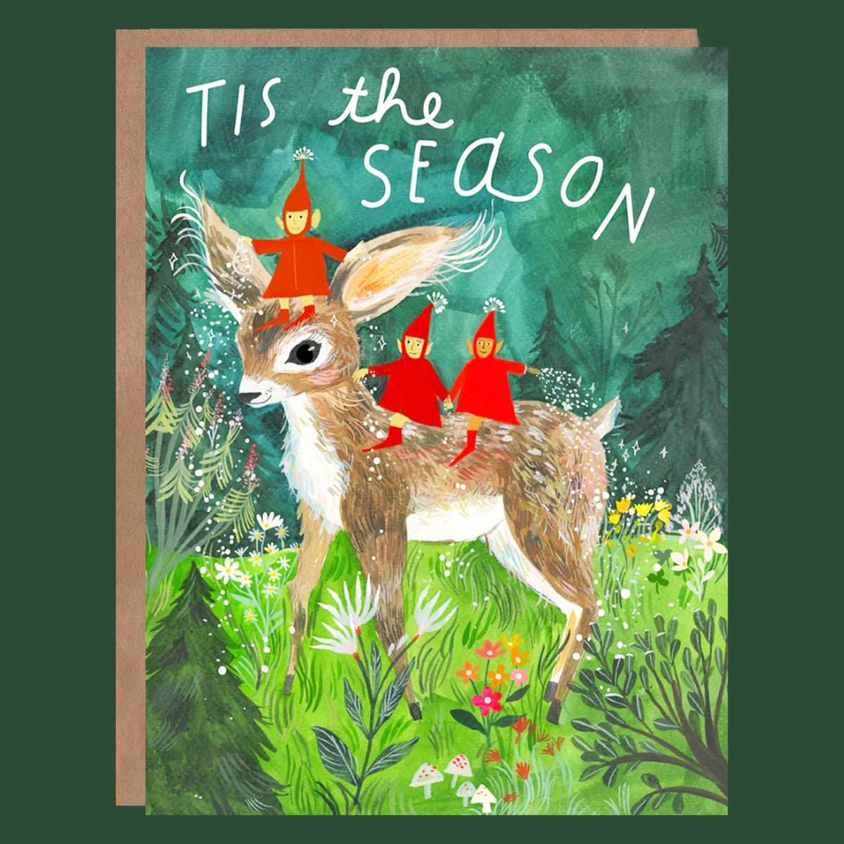 A square greeting card with a brown envelope, featuring an illustrated deer in a forest with three small figures in red outfits riding on its back. The card reads “Tis the Season” in white script, and the card is shown against a dark green background.