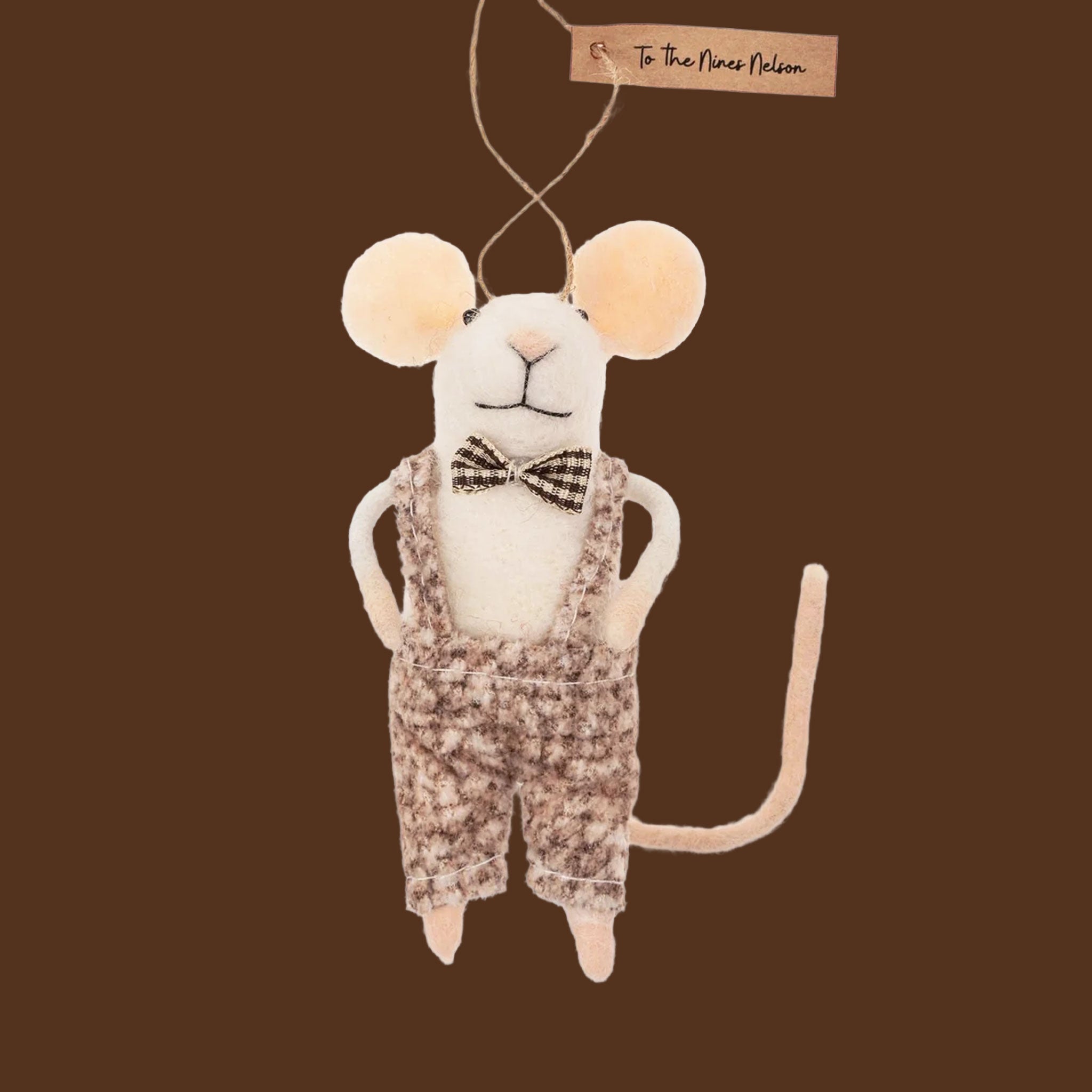 A felted mouse ornament wearing a black and white bow tie with overalls stands on a brown backtround.