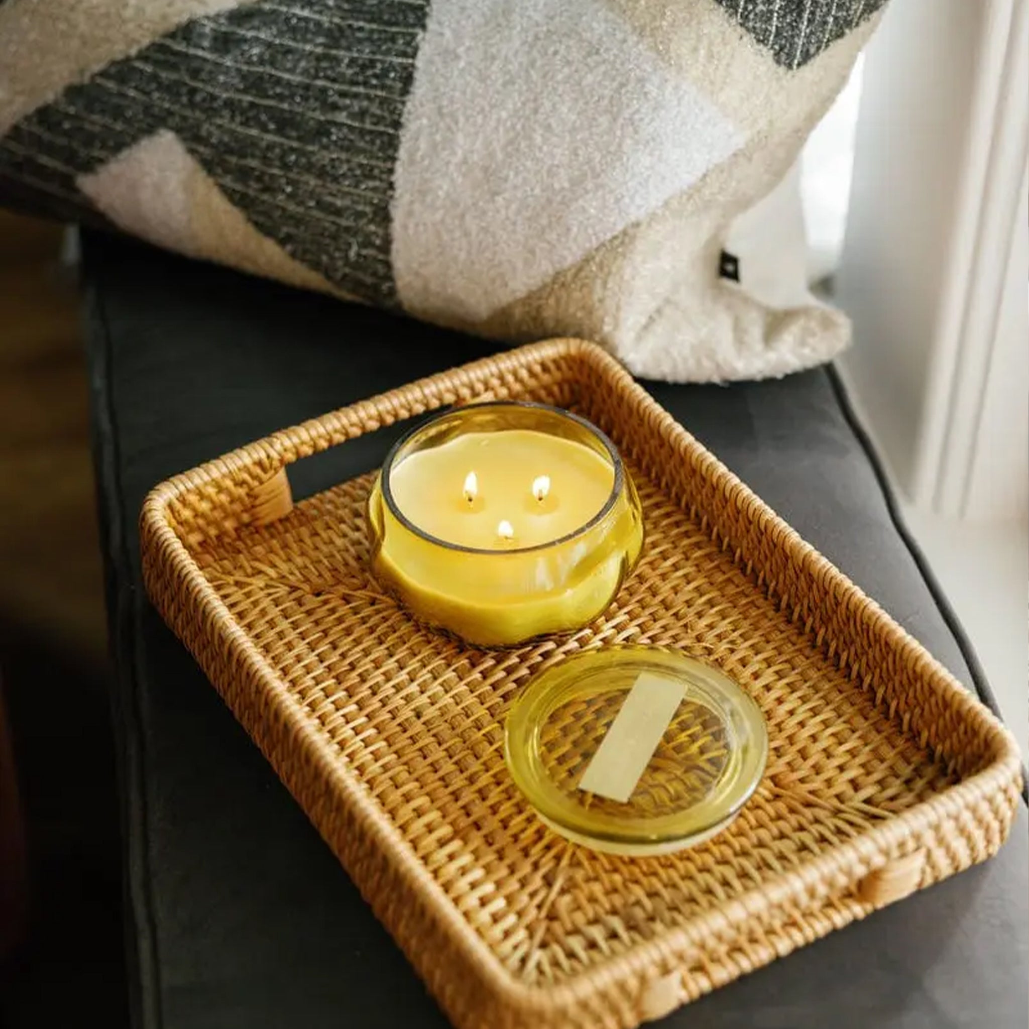 A warm, cozy scene featuring a woven tray resting on a dark sofa, holding a lit yellow three-wick candle and its lid, with a soft, textured pillow behind it.