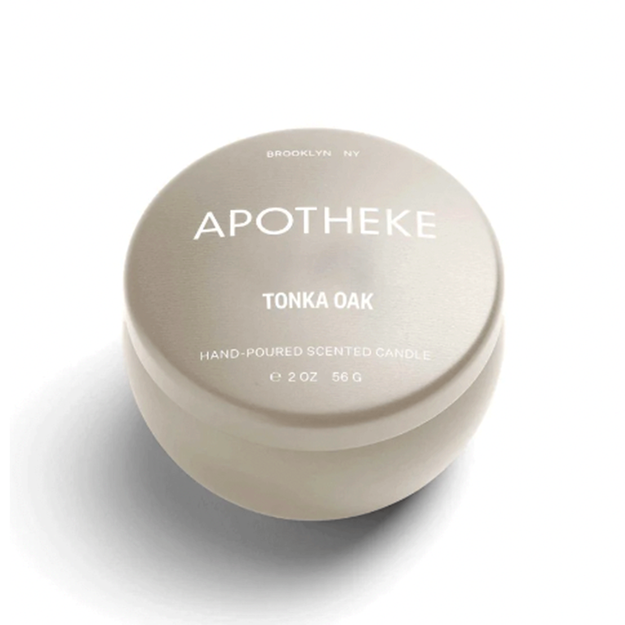On a white background is a neutral mini tin candle with white text on the lid that reads, "Apotheke Tonka Oak Hand Poured Scented Candle". 