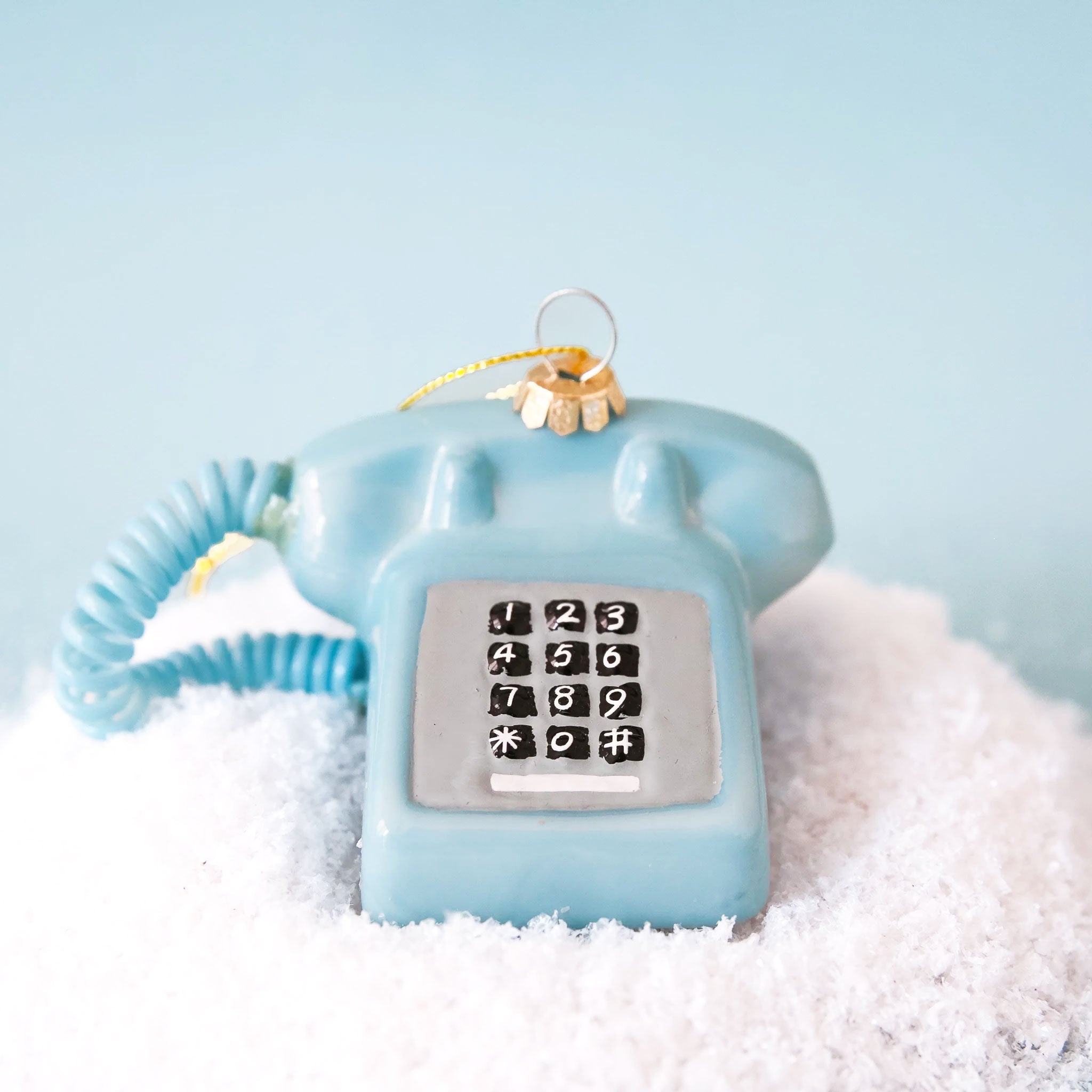 A glass touch tone phone ornament in a shiny blue shade on a snowy blue backdrop.