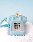 A glass touch tone phone ornament in a shiny blue shade on a snowy blue backdrop.