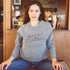 Woman with dark hair in a heather grey sweatshirt with the text "tough as a mother" in black cursive font sitting in a cozy room.
