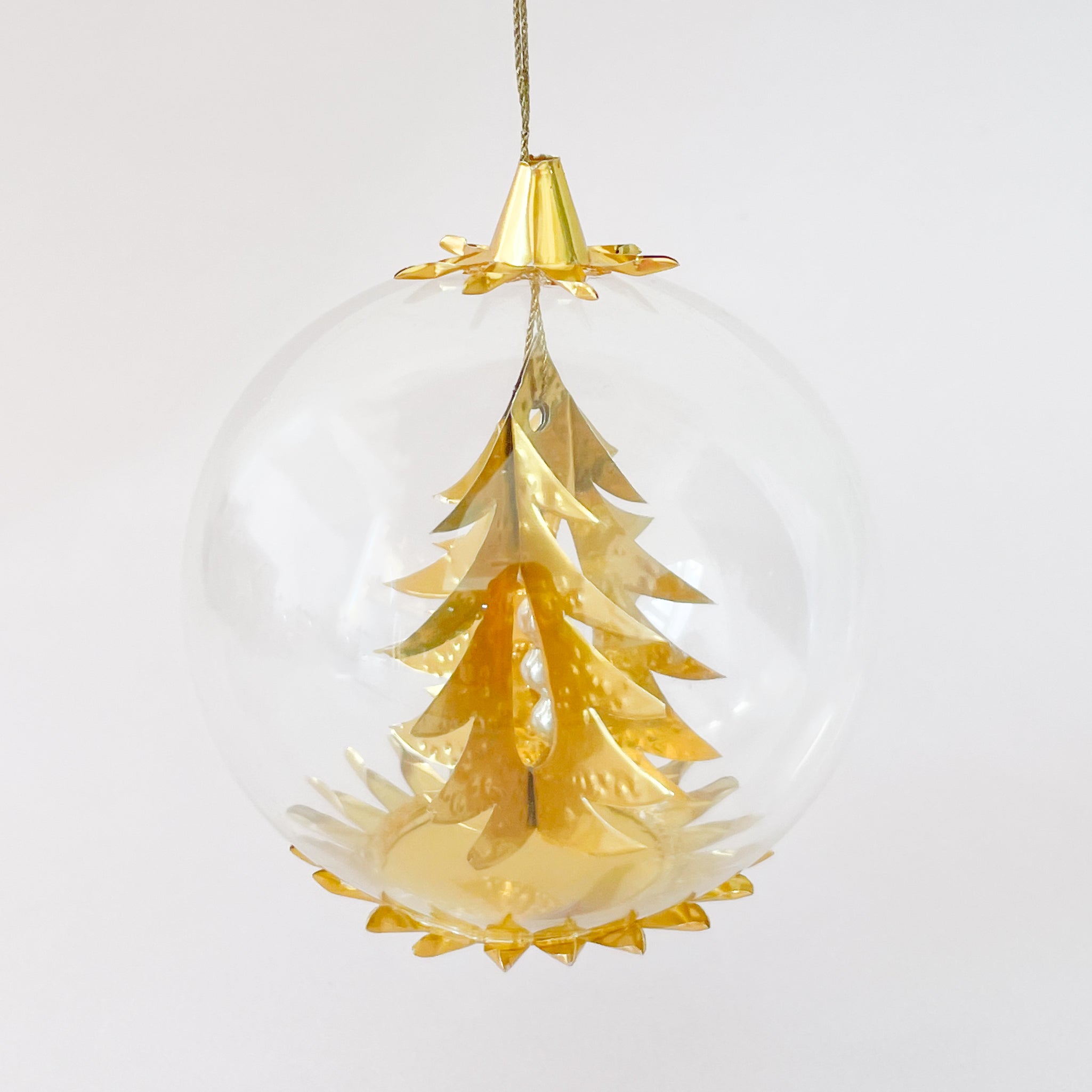 A clear round glass ornament hangs on a white background, containing a metallic gold Christmas tree with layered branches and a gold base and cap.