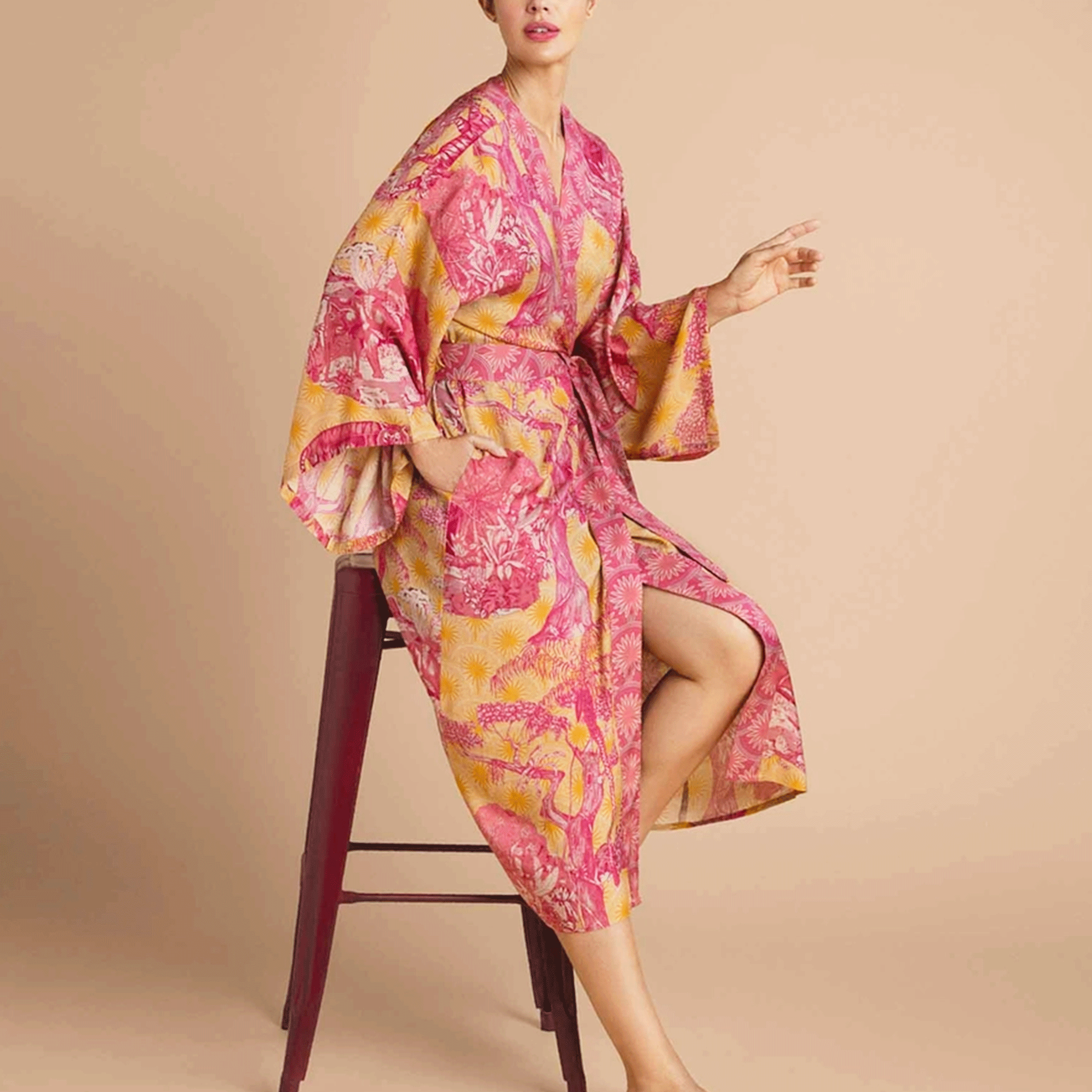 On a tan background is a model wearing a pink and yellow/orange robe with a tropical and floral print.