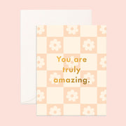 On a pink background is a daisy print checker card with gold text in the center that reads, "You are truly amazing."
