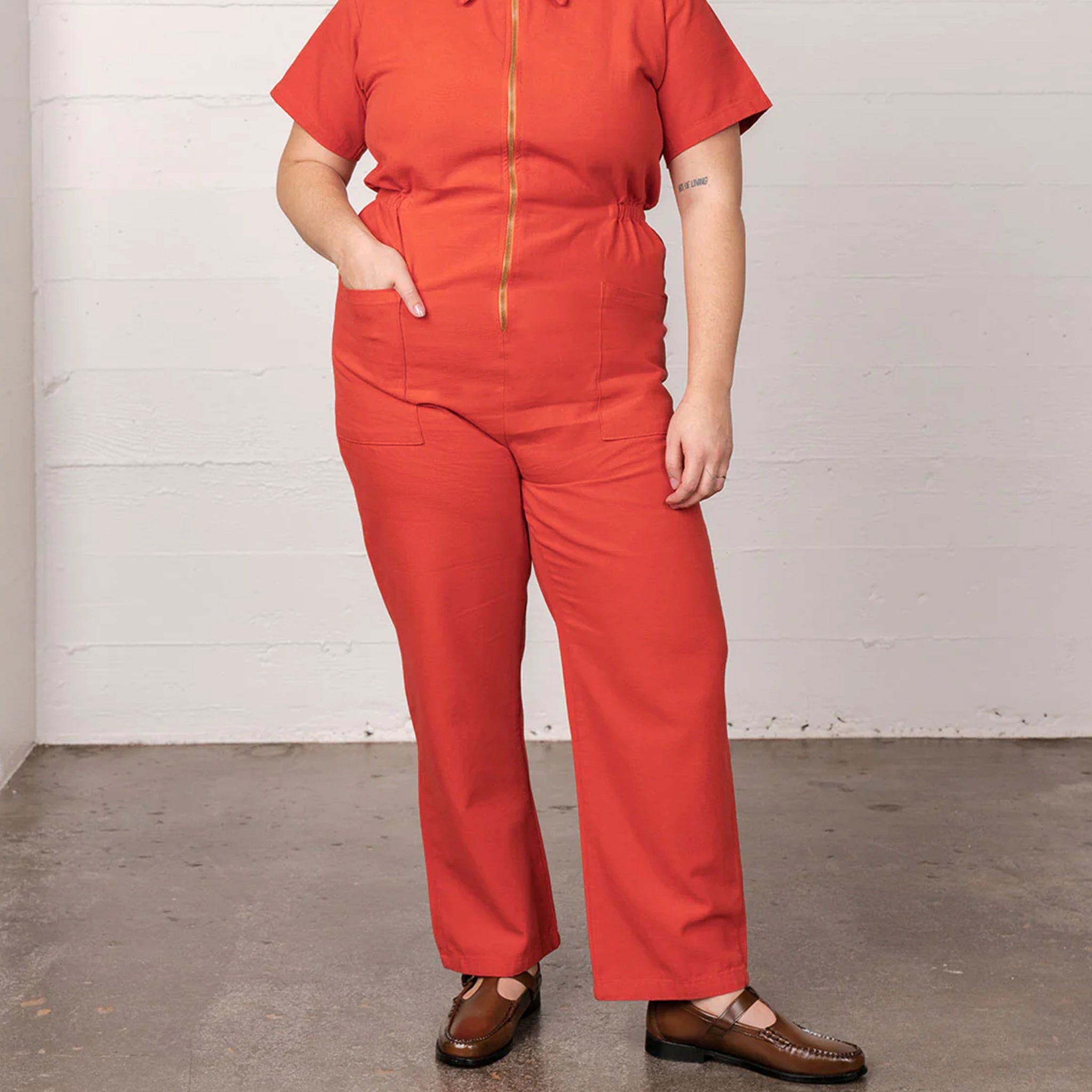 On a white background is a model wearing a short sleeve jumpsuit with a collar, side pockets and a gold zipper going down the front.