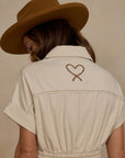 A natural colored short sleeve jumpsuit with a cinched waist and front pockets as well as a stitched rope heart detail on the back.