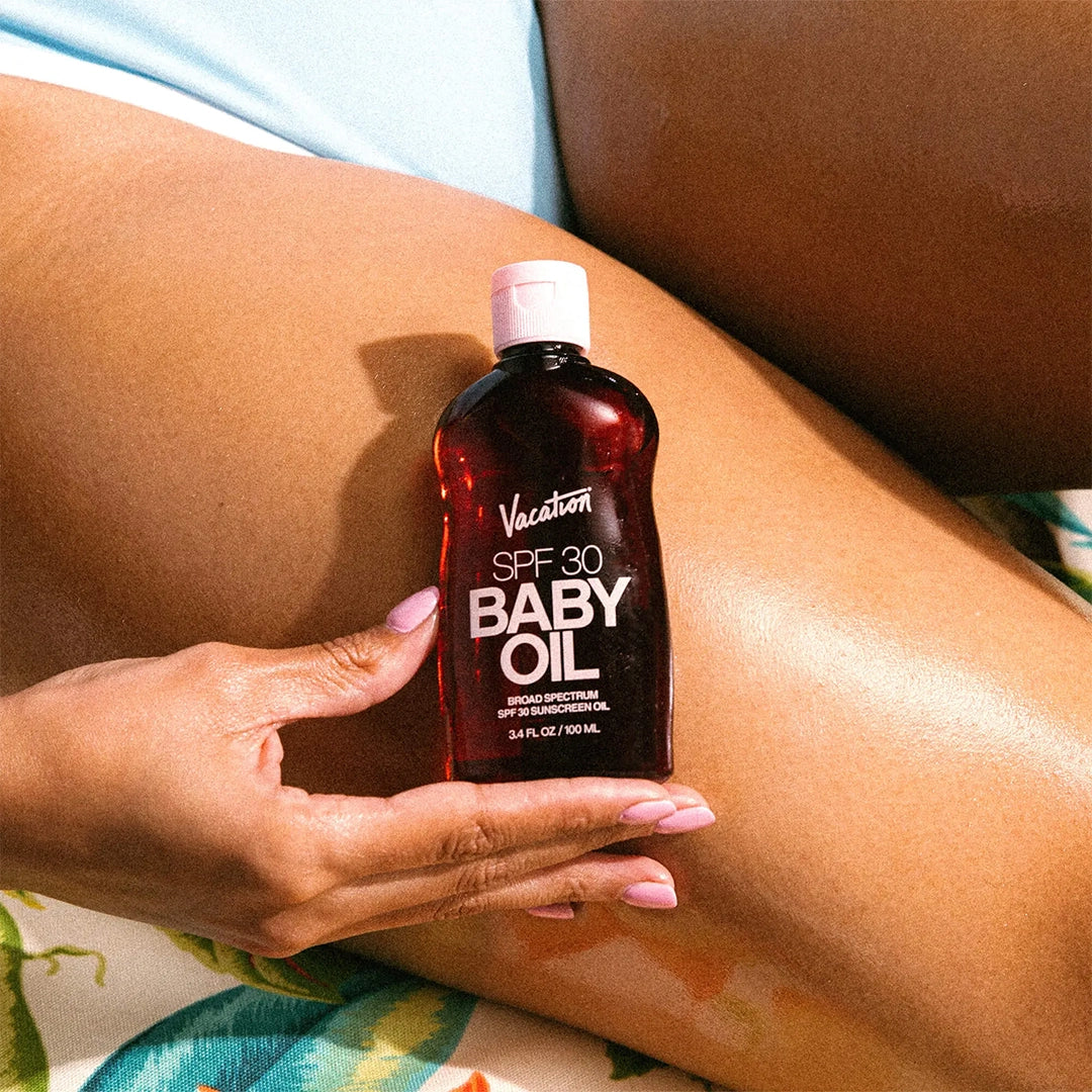 A model holding a bottle of baby oil SPF with a brownish/amber bottle and text on the front that reads, "Vacation SPF 30 Baby Oil". 
