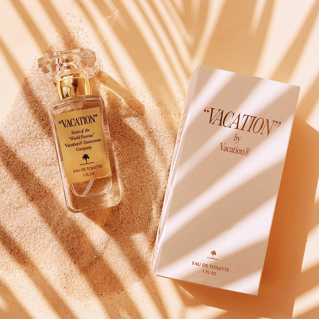 a bottle of perfume and its packaging lays on the sand with a palm leaf shadow
