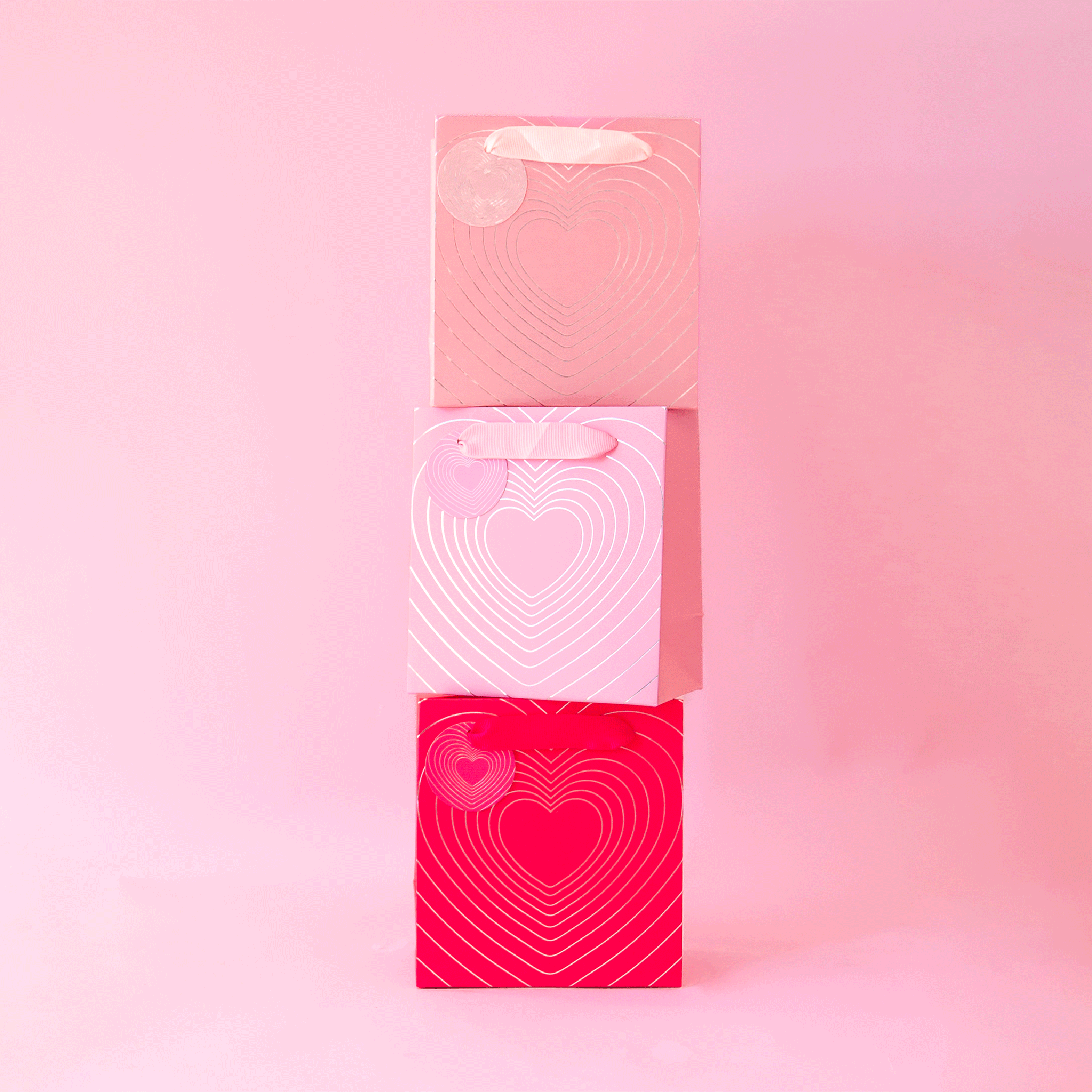 On a pink background is all three gift bag colors stacked on top of one another. 