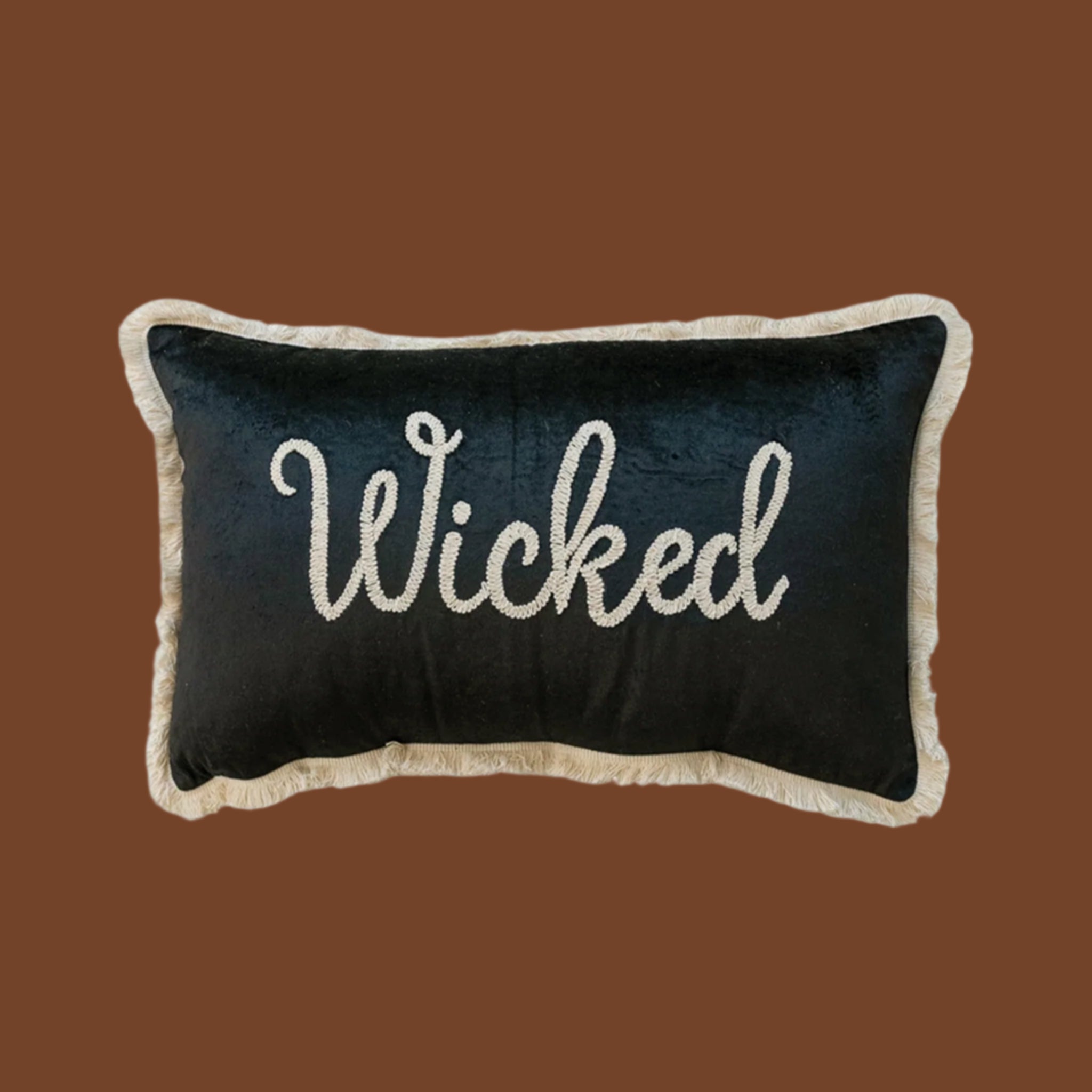 A black velvet cushion with white fringe detailing around the edge and embroidered white text in the center that reads, 'Wicked'. 