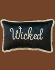 A black velvet cushion with white fringe detailing around the edge and embroidered white text in the center that reads, 'Wicked'. 