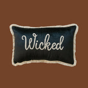 A black velvet cushion with white fringe detailing around the edge and embroidered white text in the center that reads, 'Wicked'. 