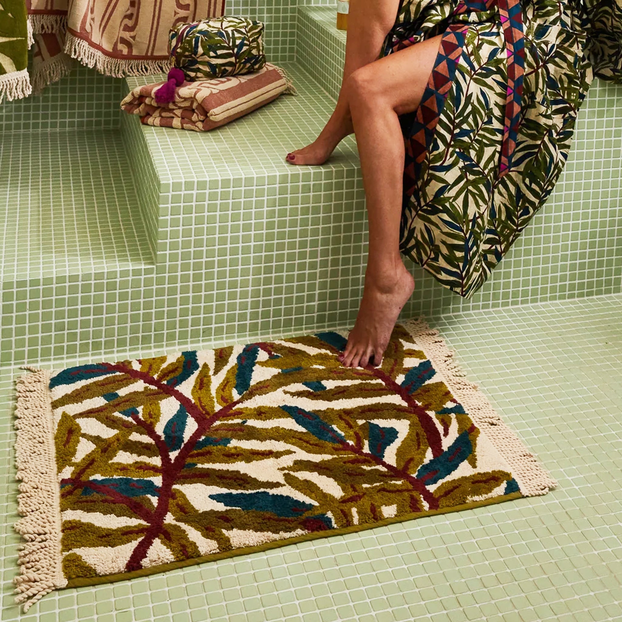 A green, teal and ivory tropical foliage printed bath mat with tassel details on each end. 