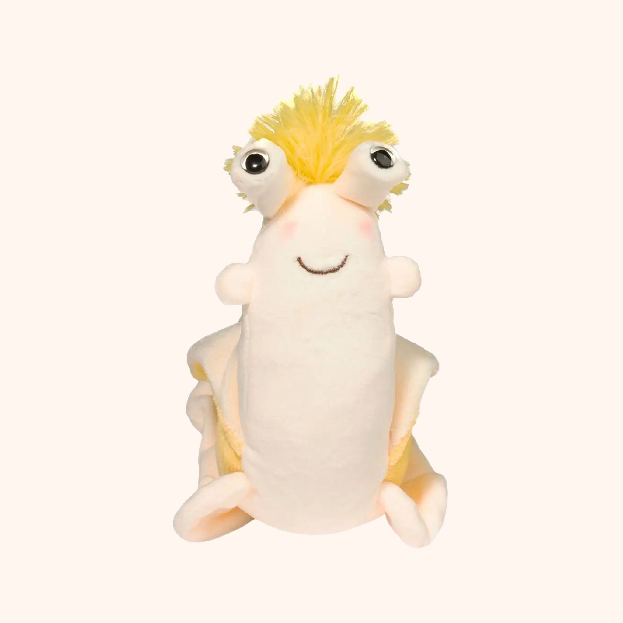 On a tan background is an ivory and bright yellow banana slug shaped stuffed animal toy with bulging eyes, a fluffy yellow pom on the top of its head. The back of its body is shaped like a banana peel.