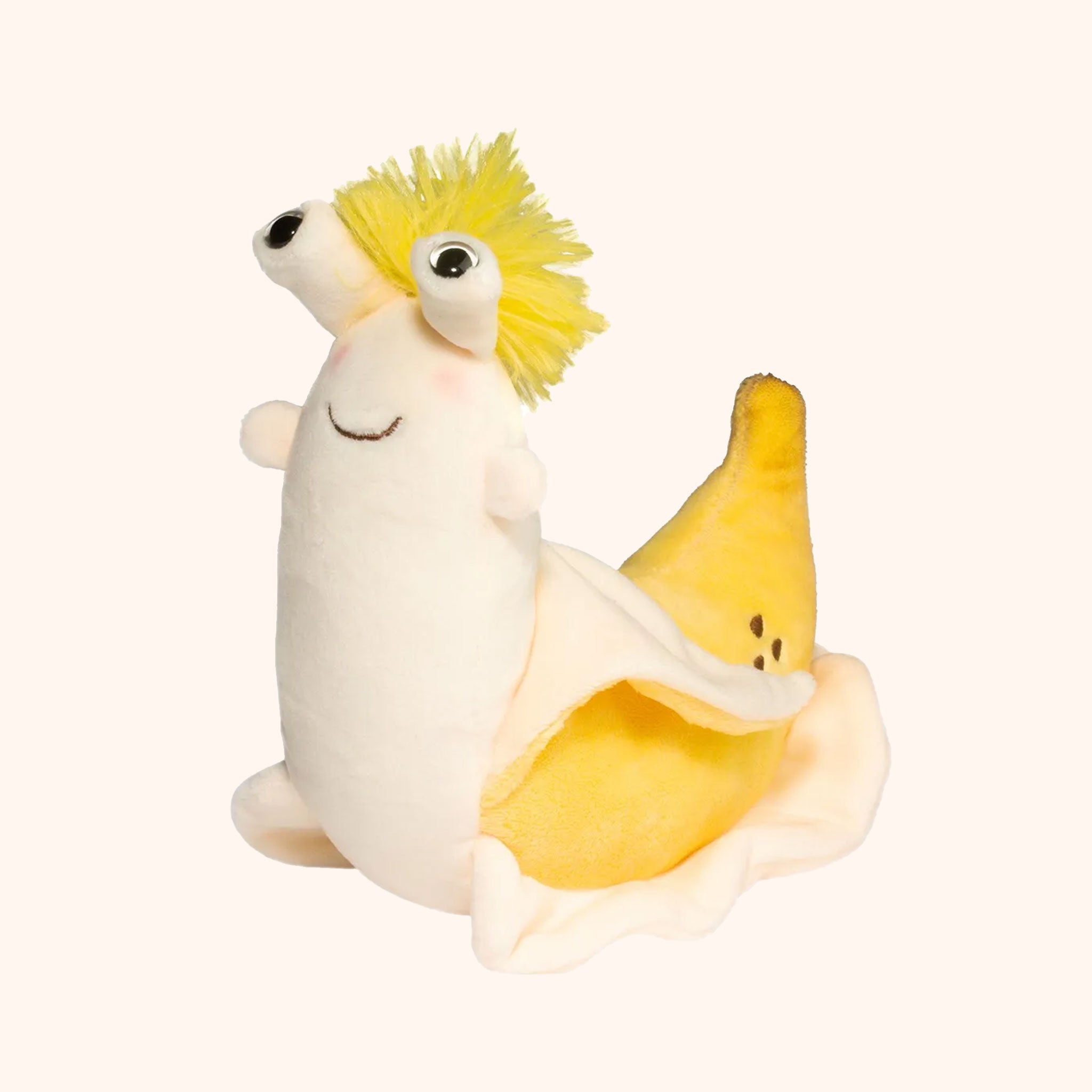 On a tan background is an ivory and bright yellow banana slug shaped stuffed animal toy with bulging eyes, a fluffy yellow pom on the top of its head. The back of its body is shaped like a banana peel. 