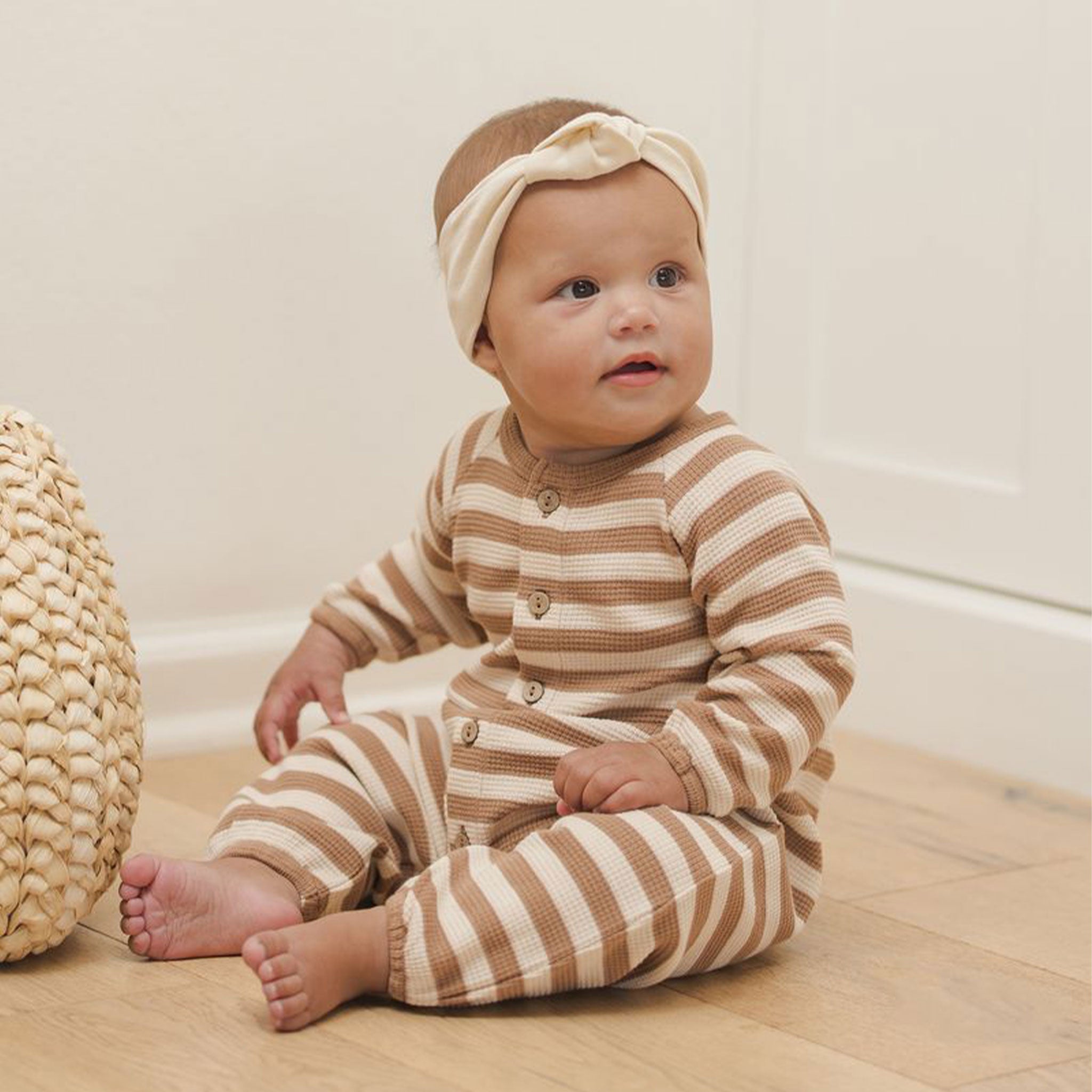 Baby wearing a long sleeve and long pant jumpsuit with a light brown and off white stripe print. The jumpsuit has 6 buttons down the front and a waffle knit texture. 