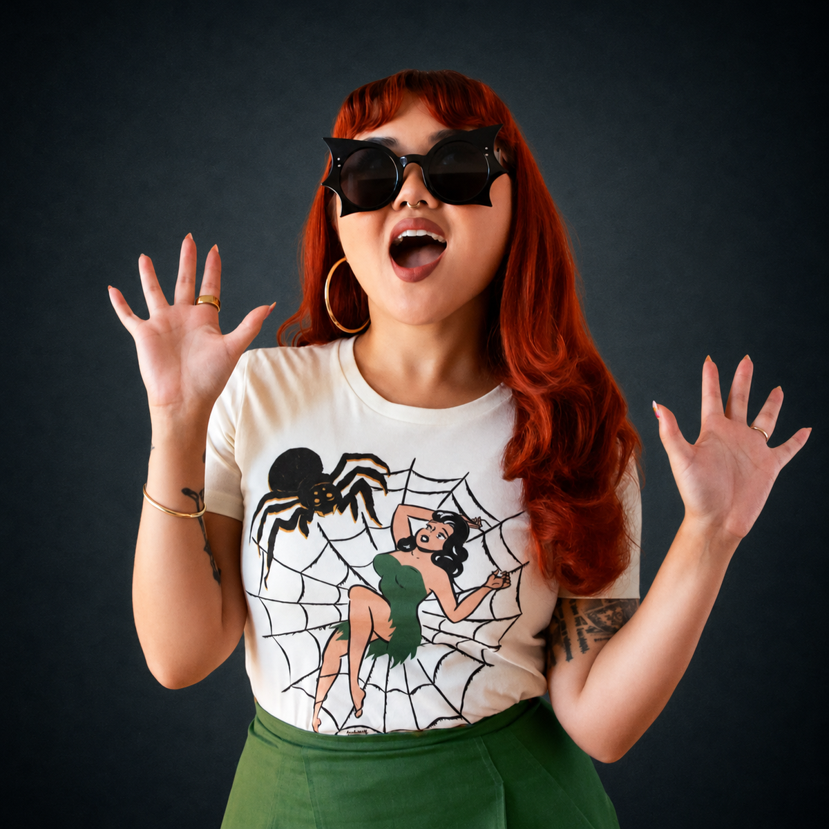 An ivory tshirt with a graphic of a woman in a green dress laying in the center of a spider web and a big spider graphic to the left. 