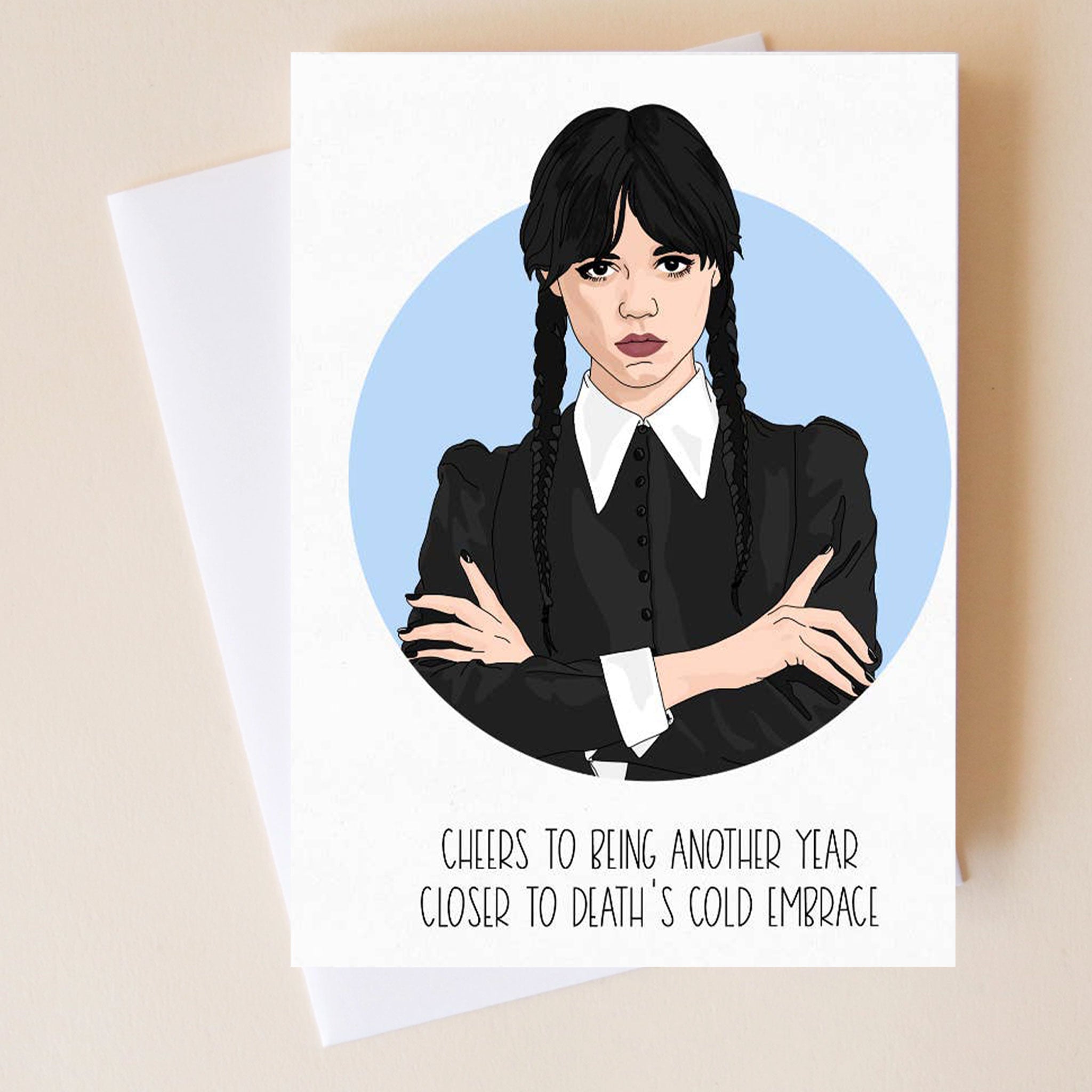 On a neutral background is a white card with an illustration of Wednesday Addams and text underneath that reads, &quot;Cheers To Being Another Year Closer To Death&#39;s Cold Embrace&quot;. 