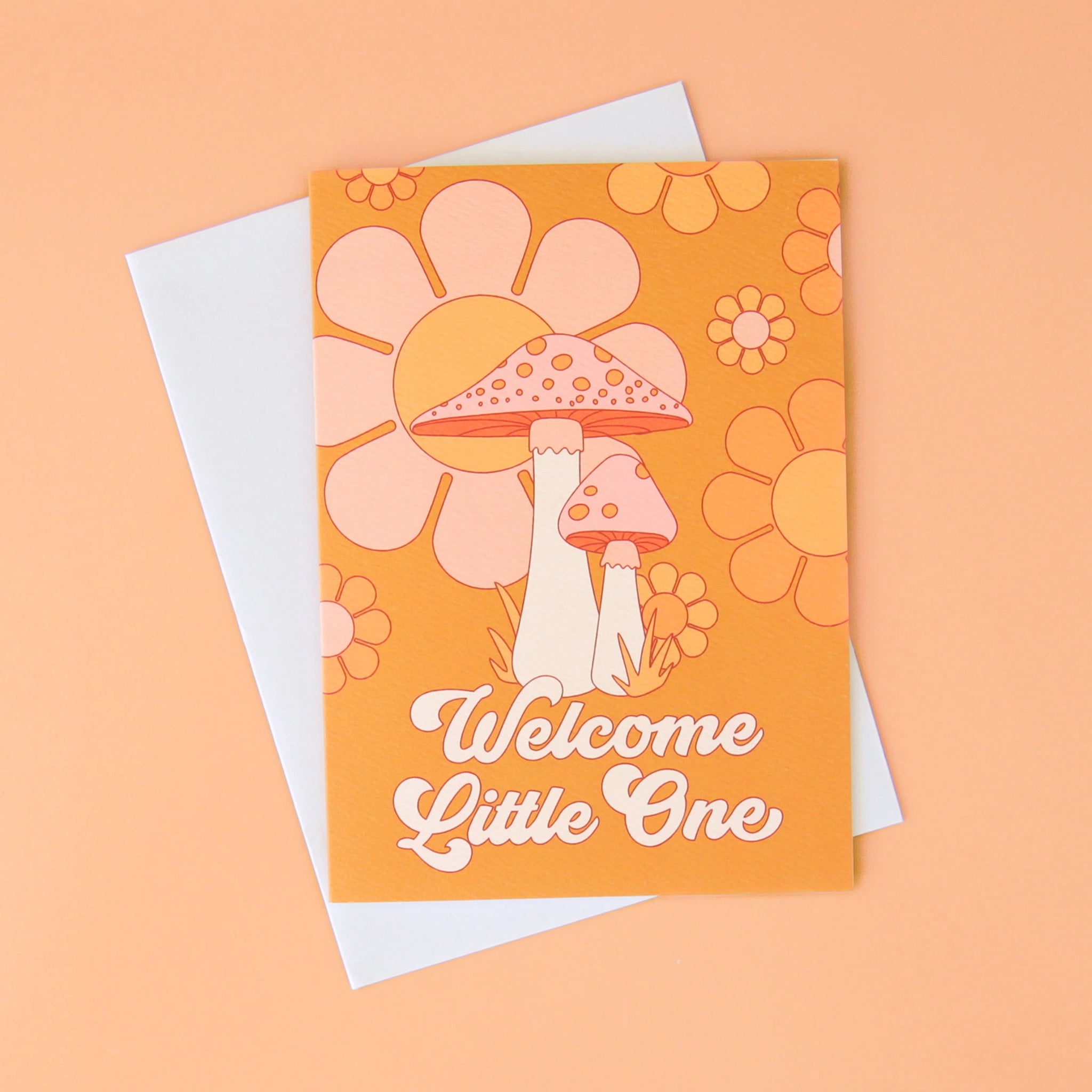 On a peachy background is an orange card with a floral and mushroom design along with white text that reads, &quot;Welcome Little One&quot;.