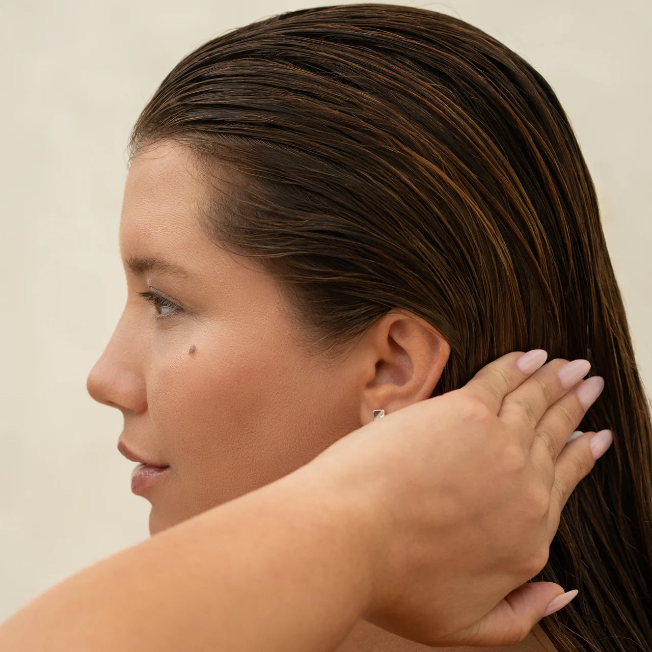 Side profile of a woman with wet, slicked-back hair, using her hand to smooth it behind her ear against a neutral background.