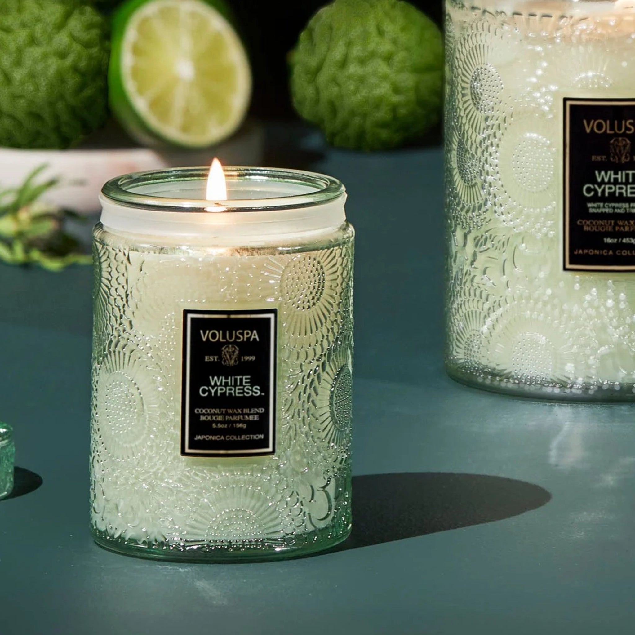 A green glass decorative candle with a single wick and black rectangle label in front that reads, &quot;Voluspa White Cypress&quot;. 