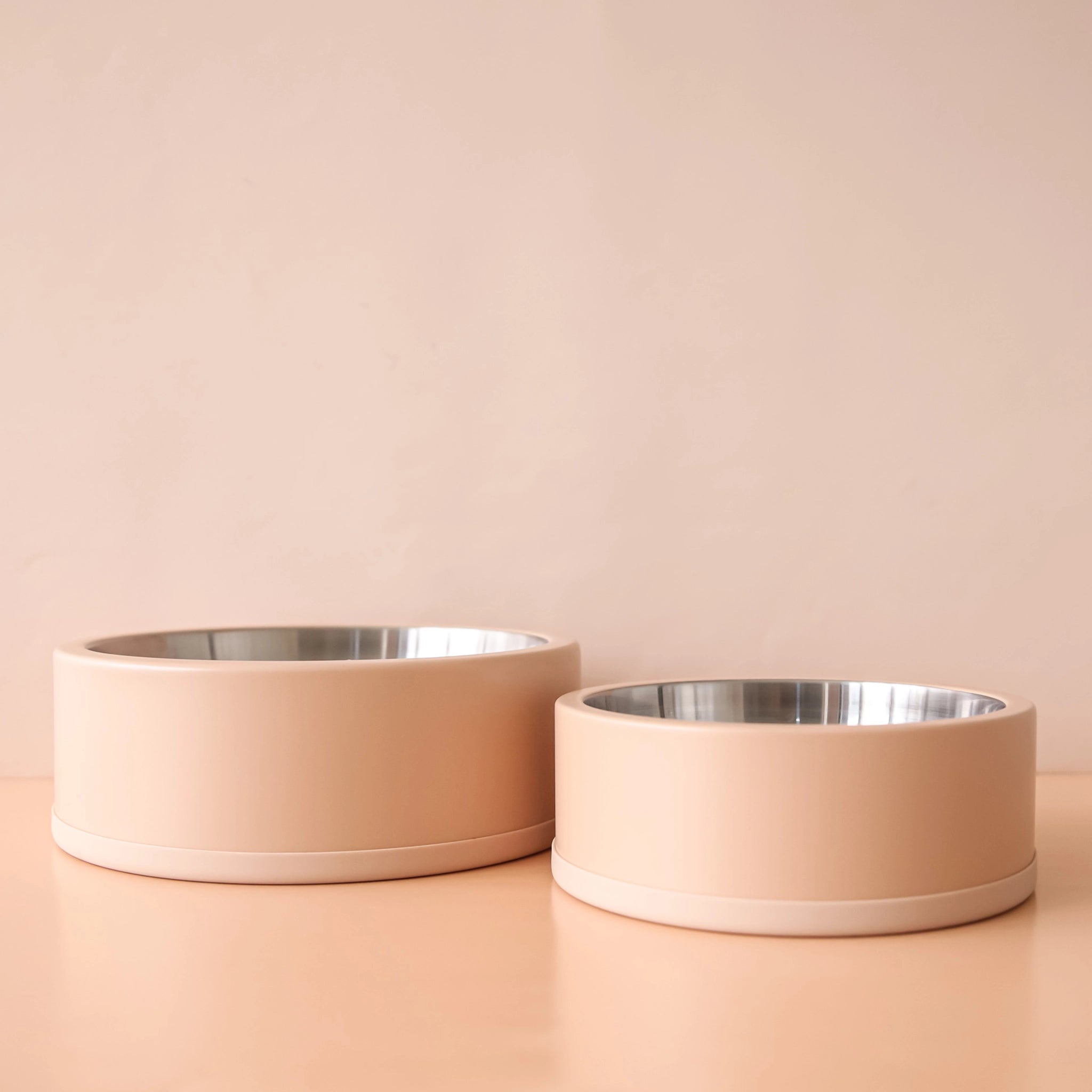 On a peachy background is two different sized dog bowls with stainless steel interiors and a tan exterior.