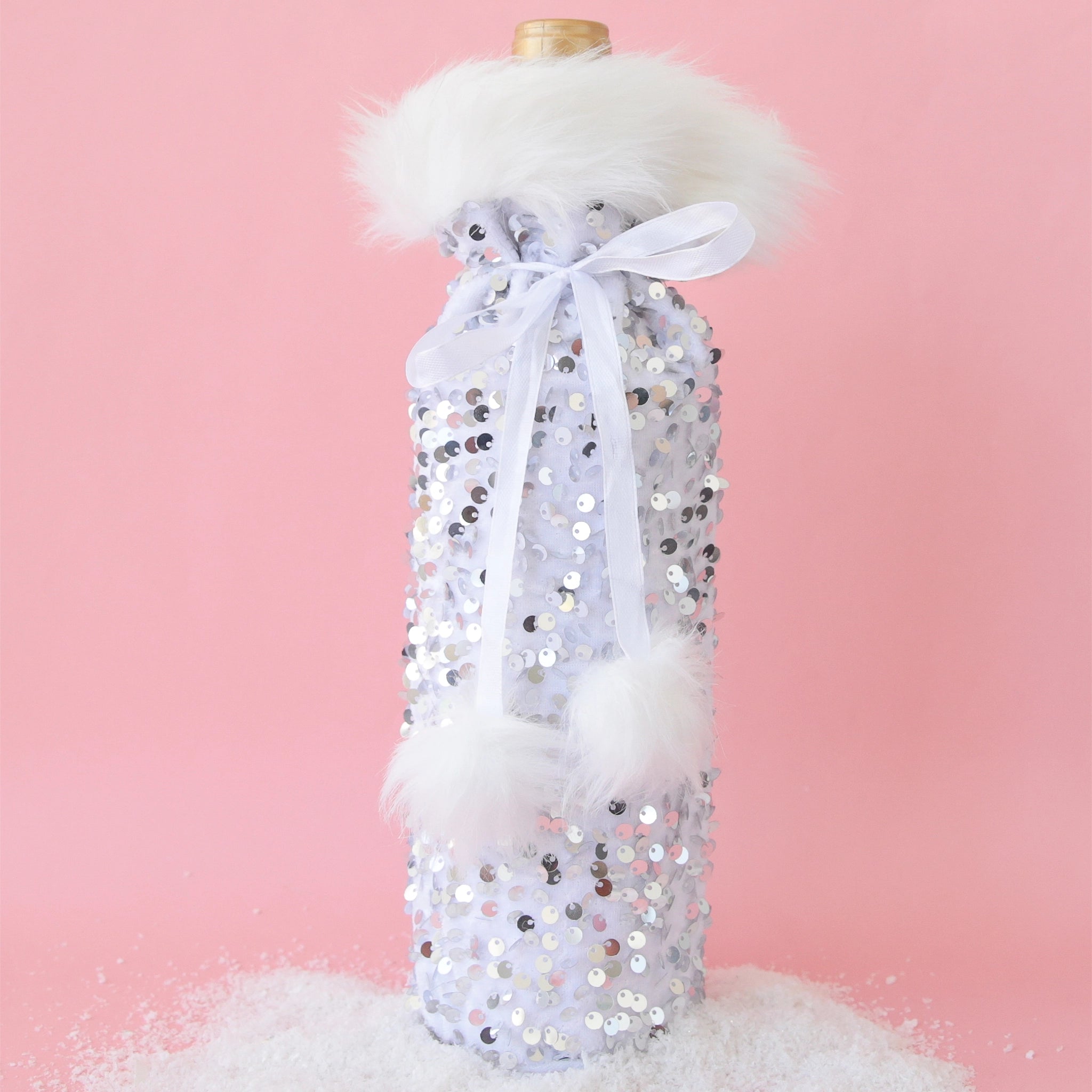 On a pink background is a silver sequin wine gift bag with white faux fur around the opening and a white pom pom bow around the top. 