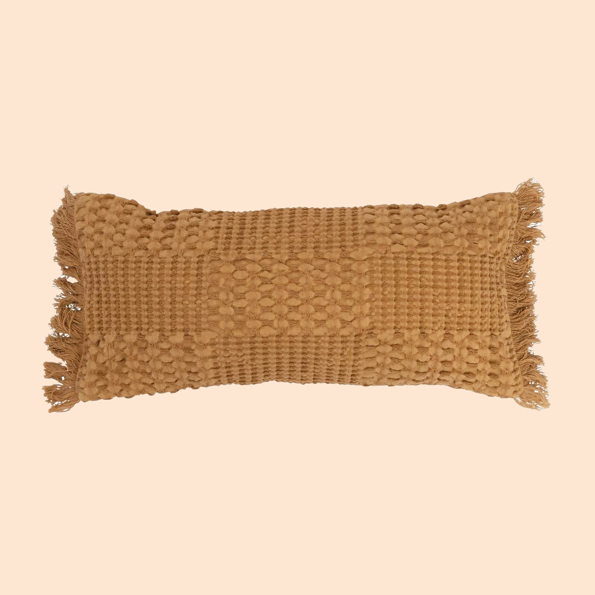 A toffee colored woven lumbar pillow with fringe details on each end. 