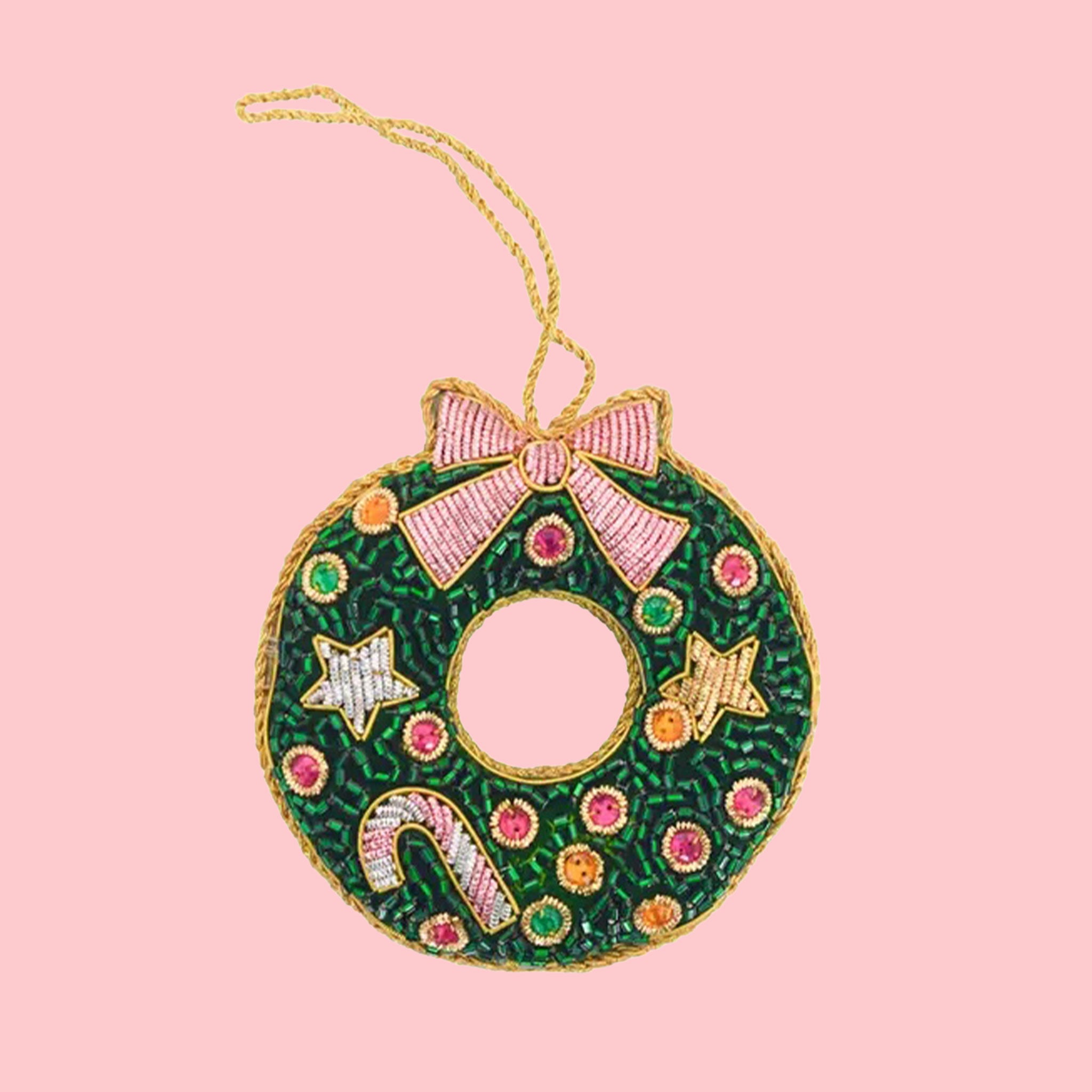 A beaded green wreath ornament with a pink bow, colorful jewel-like dots, star accents, and a small candy cane, hanging from a gold cord against a pale pink background.