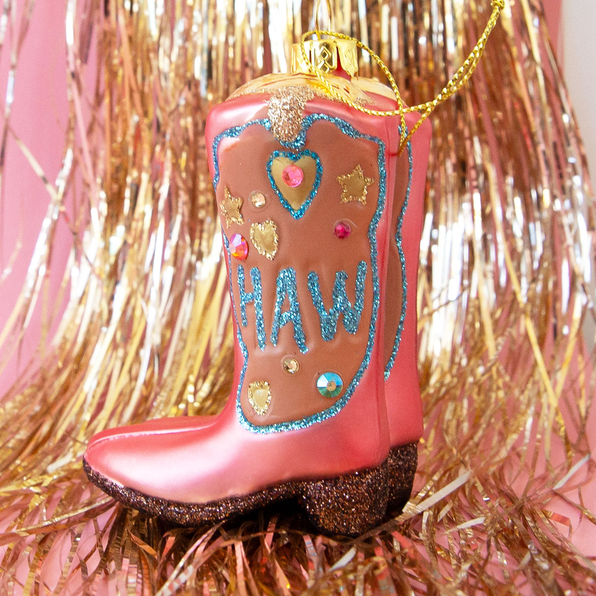 On a pink and gold tinsel background is a glass ornament in the shape of pink cowgirl boots with blue pink and gold detailing along with glitter text that reads, &quot;Yee&quot; on one side and &quot;Haw&quot; on the other. 