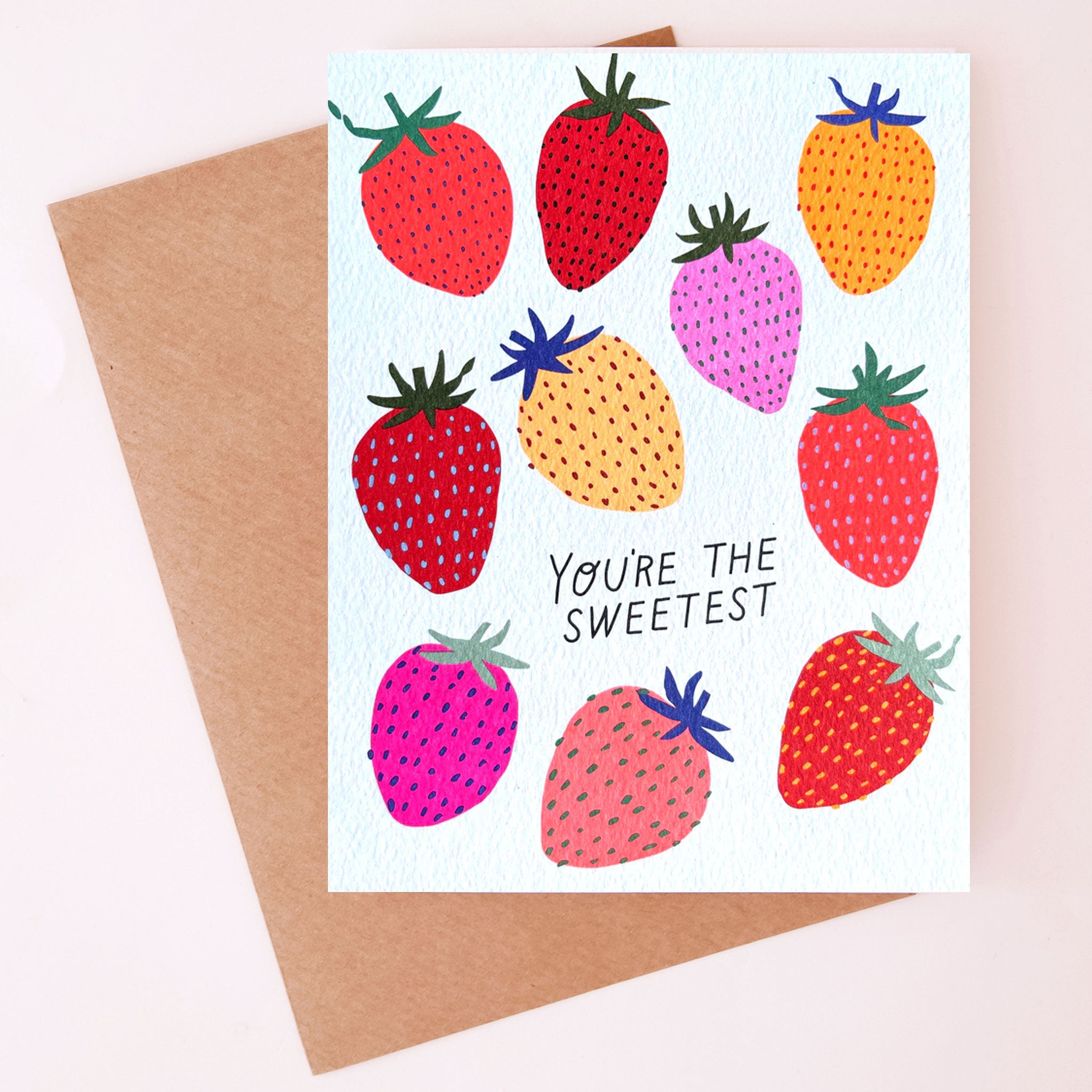 On a neutral background is a white greeting card with various strawberry prints on the front in shades of red, pink and yellow along with text in the center that reads, "You're The Sweetest". 
