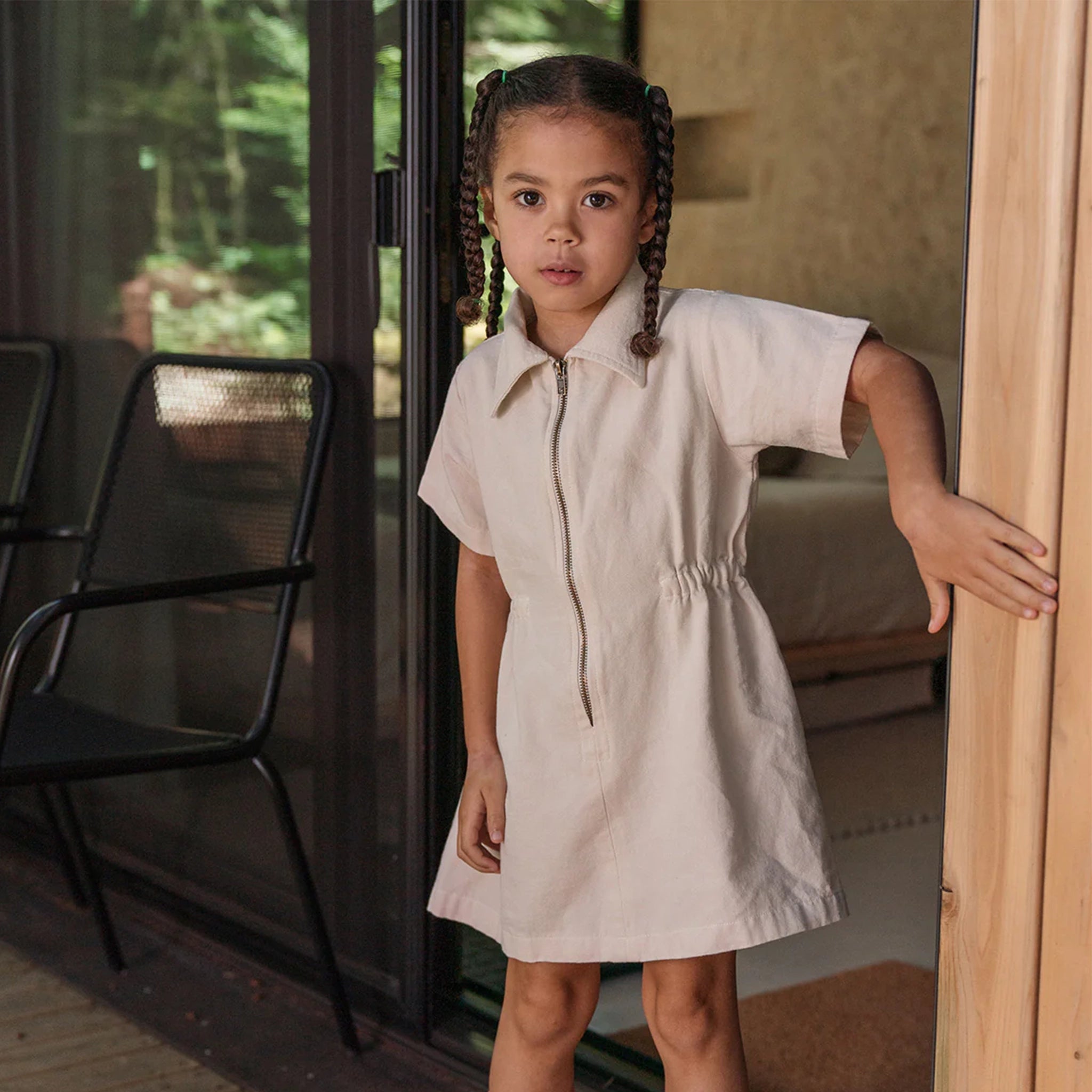 A children&#39;s model wearing a neutral tan utility dress with a collar and a zip up the front. 