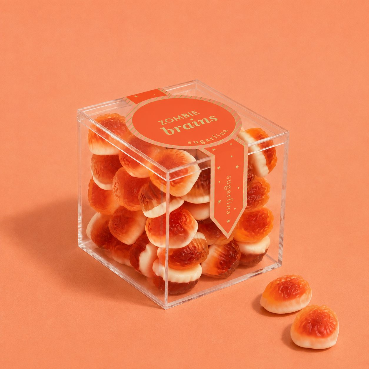 A clear square plastic box filled with small zombie brain gummy candies, with two candies placed outside the box on a peach-orange background. The box has an orange label that reads 'zombie brains' and 'sugarfina'.