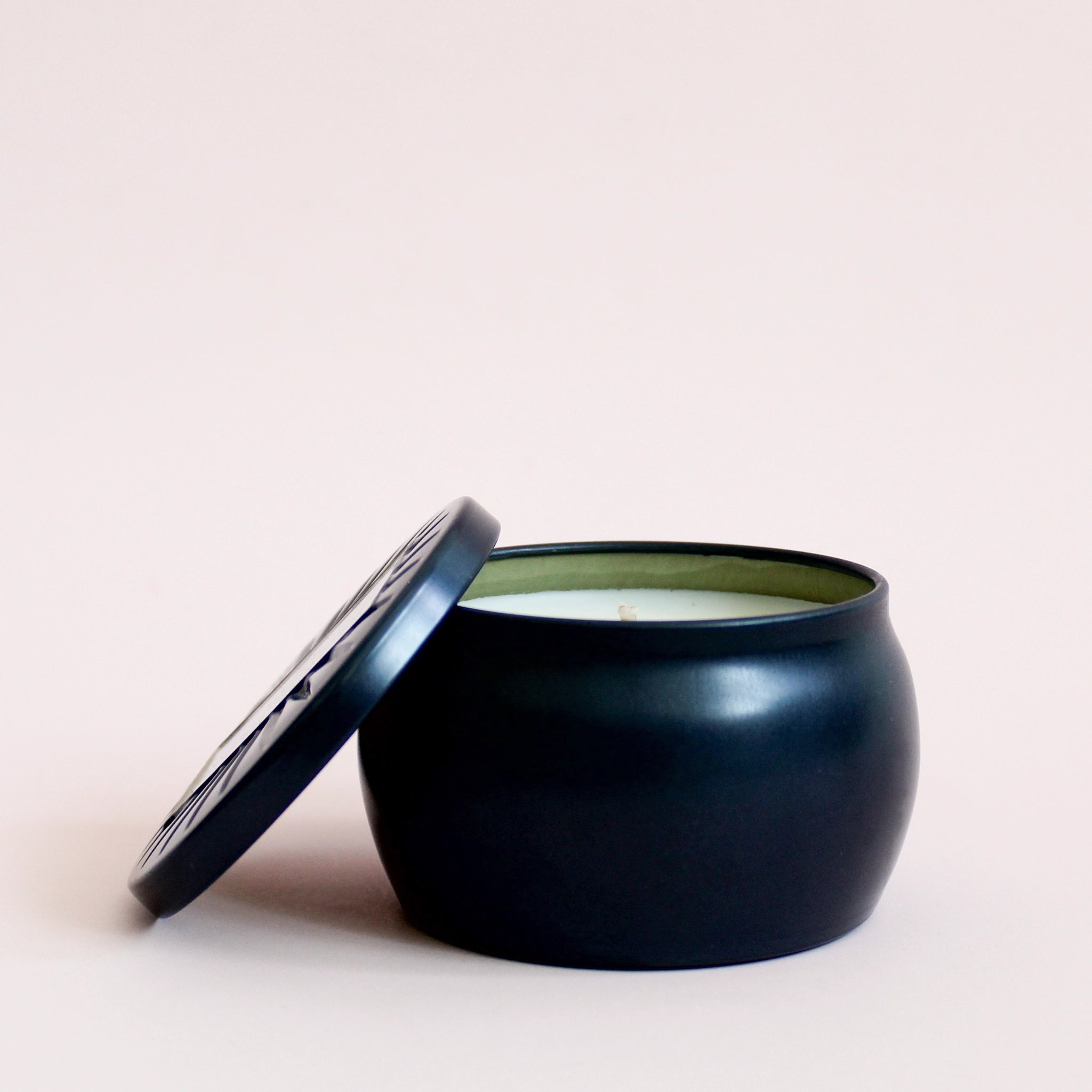 On a cream background is a round tin single wick candle in a navy blue shade along with a label on the lid that reads, "Voluspa Makassar Ebony & Peach".