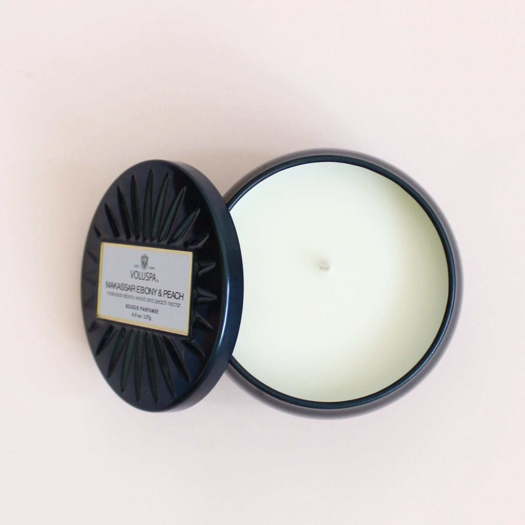 On a cream background is a round tin single wick candle in a navy blue shade along with a label on the lid that reads, "Voluspa Makassar Ebony & Peach".