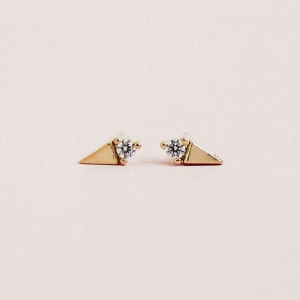 In front of a soft pink background is a pair stud earrings. On one side of each stud is a gold triangle and on the other side is a white diamond. The diamond is on the flat side of the triangle. 