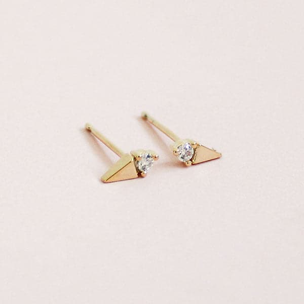 In front of a soft pink background is a pair stud earrings. On one side of each stud is a gold triangle and on the other side is a white diamond. The diamond is on the flat side of the triangle. 